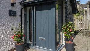 A building with a dark grey door, a sign reading "ROSE HOUSE", and two potted plants with pink flowers. - Rose House (Windermere)