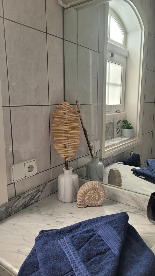 Un baño con un espejo grande, un florero con una rama seca, una concha y una toalla en la encimera.