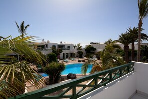 A pool area with a clear blue pool, surrounded by palm trees and white buildings with balconies. - Casa Rocas Negras (Tías)