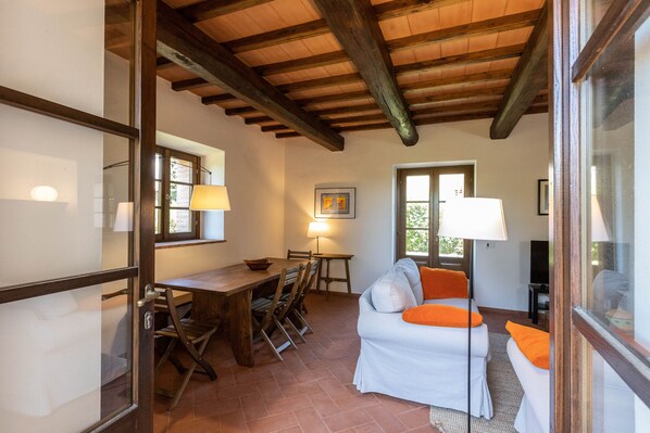 A cozy room with a wooden ceiling, a dining table with chairs, a sofa with orange cushions, and a lamp. - Sweet country home (Montorgiali)