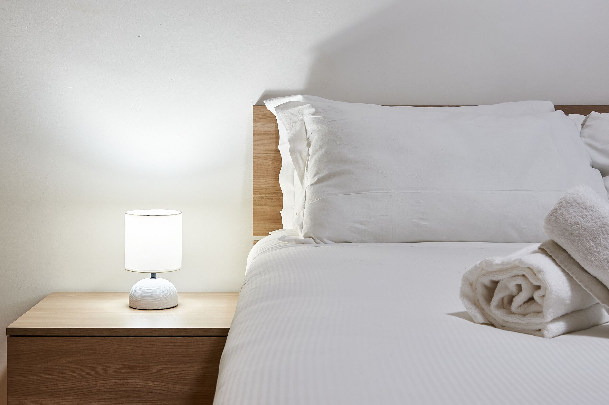 A neatly made bed with white linens, a rolled towel, and a bedside lamp on a wooden nightstand.