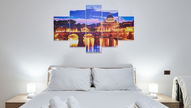A hotel room with a large bed, white bedding, two bedside lamps, and a wall-mounted artwork of a cityscape. - Anastasio II apartment  (Rome)