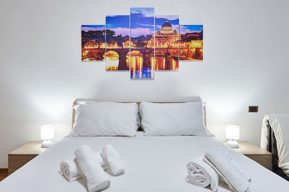 A hotel room with a large bed, white bedding, two bedside lamps, and a wall-mounted artwork of a cityscape.