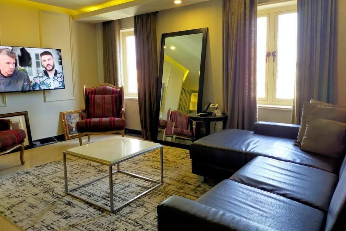 A modern hotel room with a sofa, armchair, and a television displaying two people.