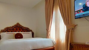 A hotel room with a wooden headboard, a bed with white linens, a television on the wall, and a window with curtains. - Catherine Villa (Lagos)