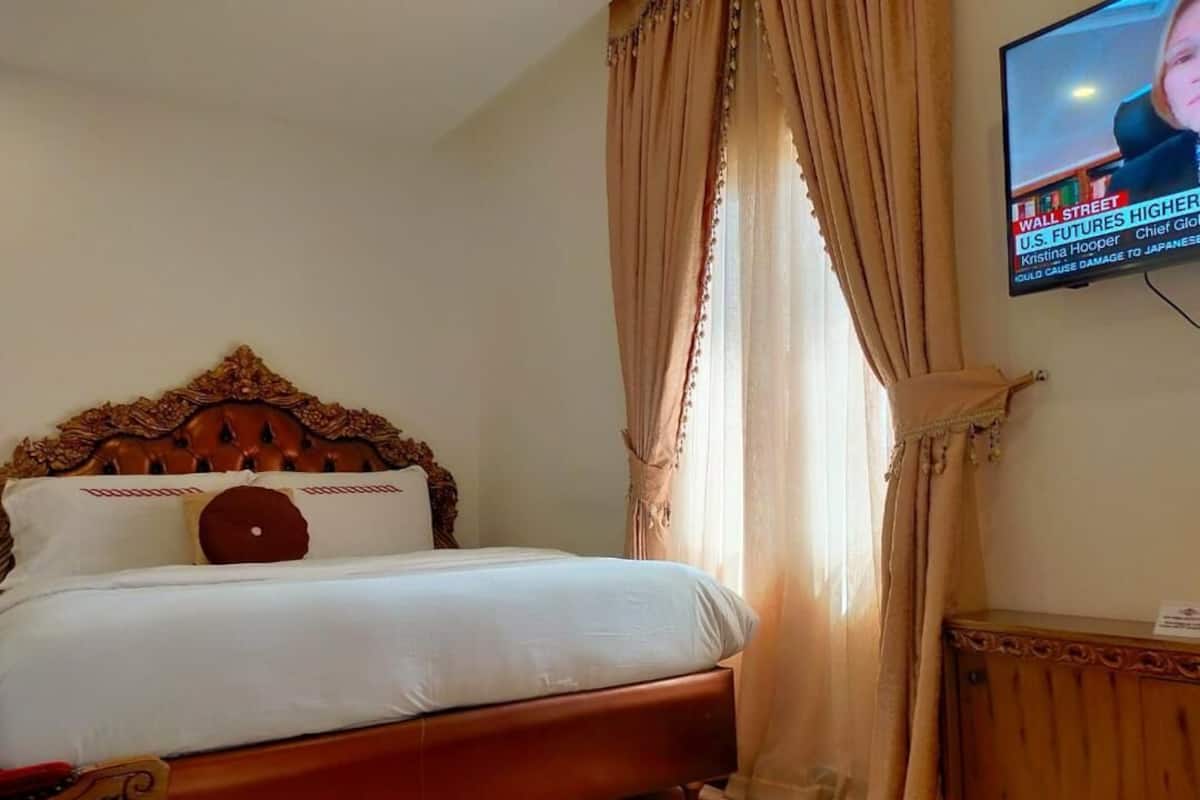 A hotel room with a wooden headboard, a bed with white linens, a television on the wall, and a window with curtains.