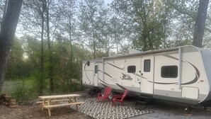 A recreational vehicle (RV) parked in a wooded area with a picnic table and red chairs.