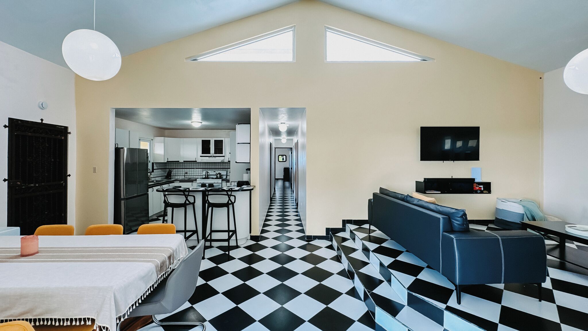 Une chambre d’hôtel moderne avec un sol à carreaux, un coin repas avec des tabourets de bar, un canapé et un téléviseur fixé au mur.