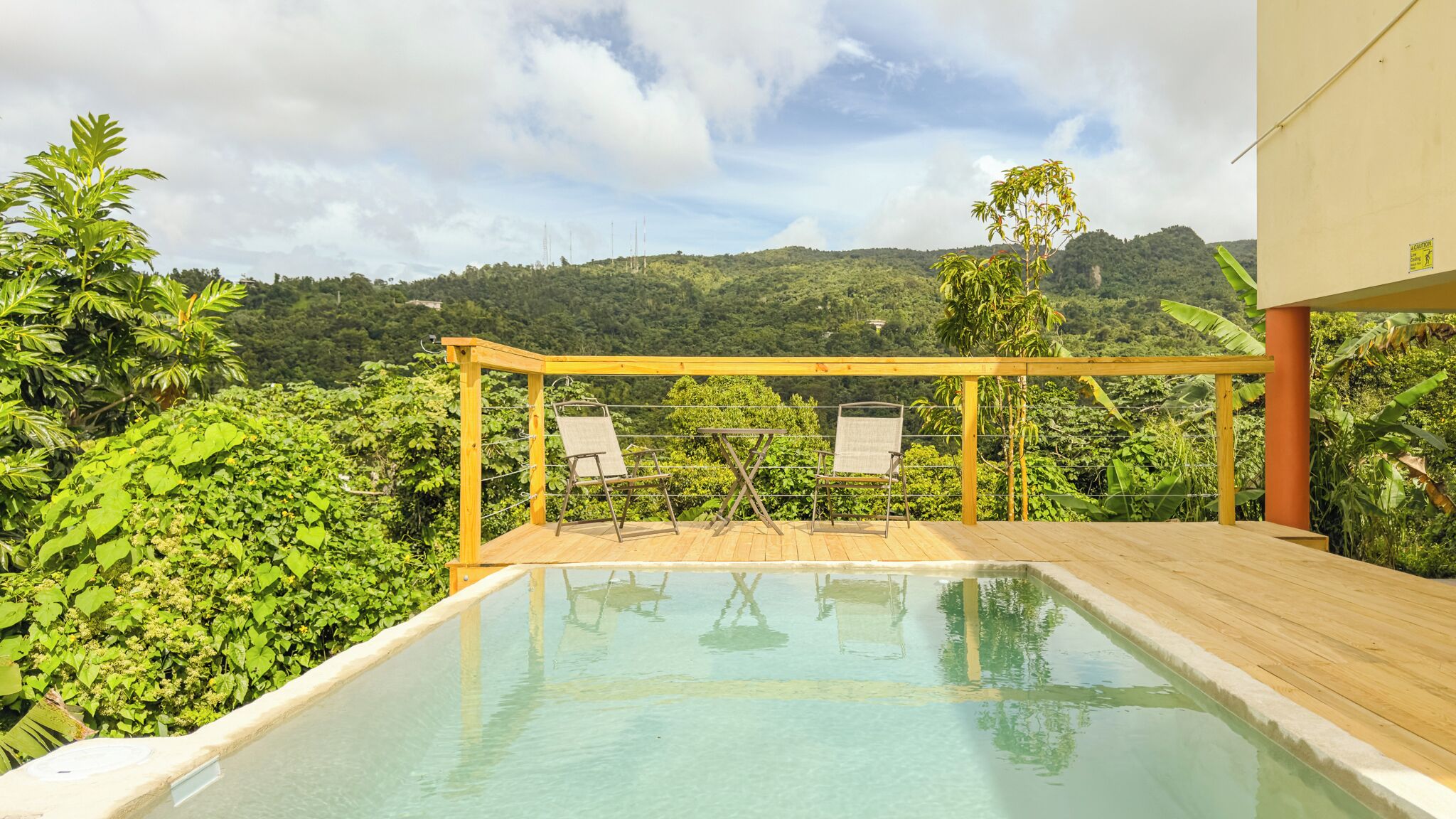 A pool with two chairs and a wooden pergola, surrounded by lush greenery and a mountainous backdrop.