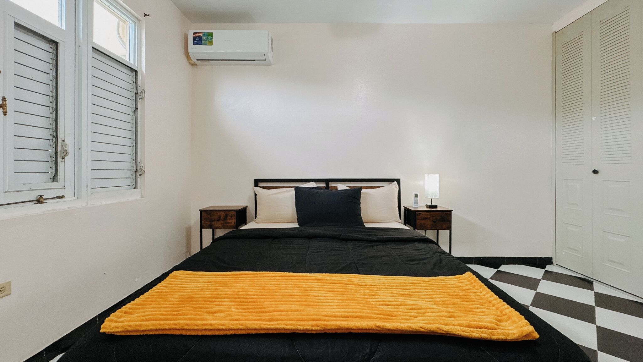 Una habitación pequeña y limpia con cama, mesita de noche, lámpara y ventana con persianas.