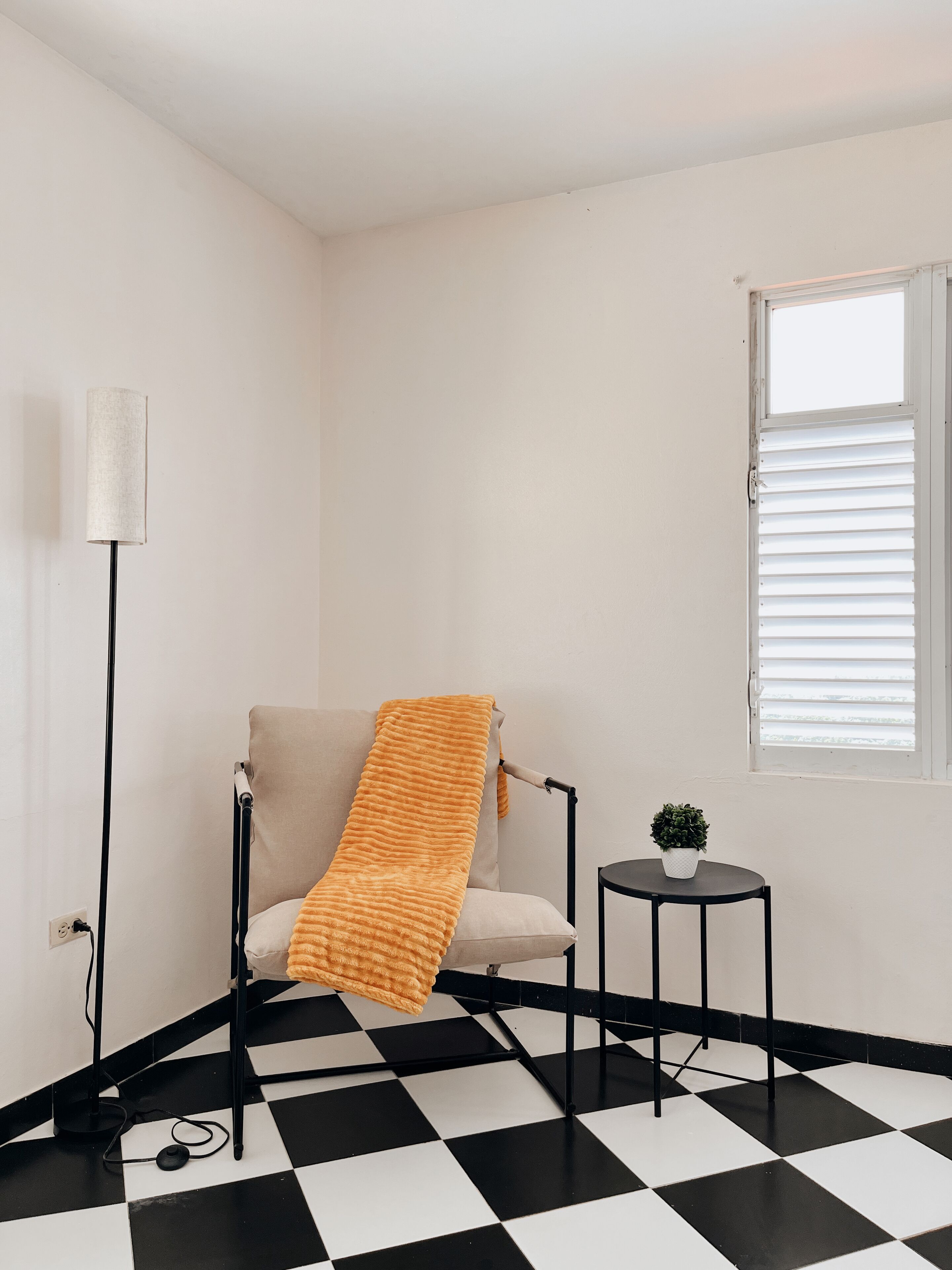 A room with a checkered floor, a chair with a blanket, a small table with a plant, a floor lamp, and a window with blinds.