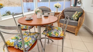 A sunlit patio with wicker furniture, a round wooden table, and a view of a brick house through large windows.