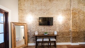 A dining area with a brick wall, a wooden table with two chairs, and a flat-screen TV mounted above the table. - Experience Mississippi Art at 'The Artistry' (Laurel)