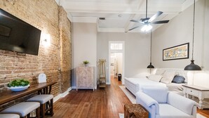 A modern living room with a brick wall, wooden flooring, a white sofa, a coffee table, and a TV mounted on the wall. - Experience Mississippi Art at 'The Artistry' (Laurel)