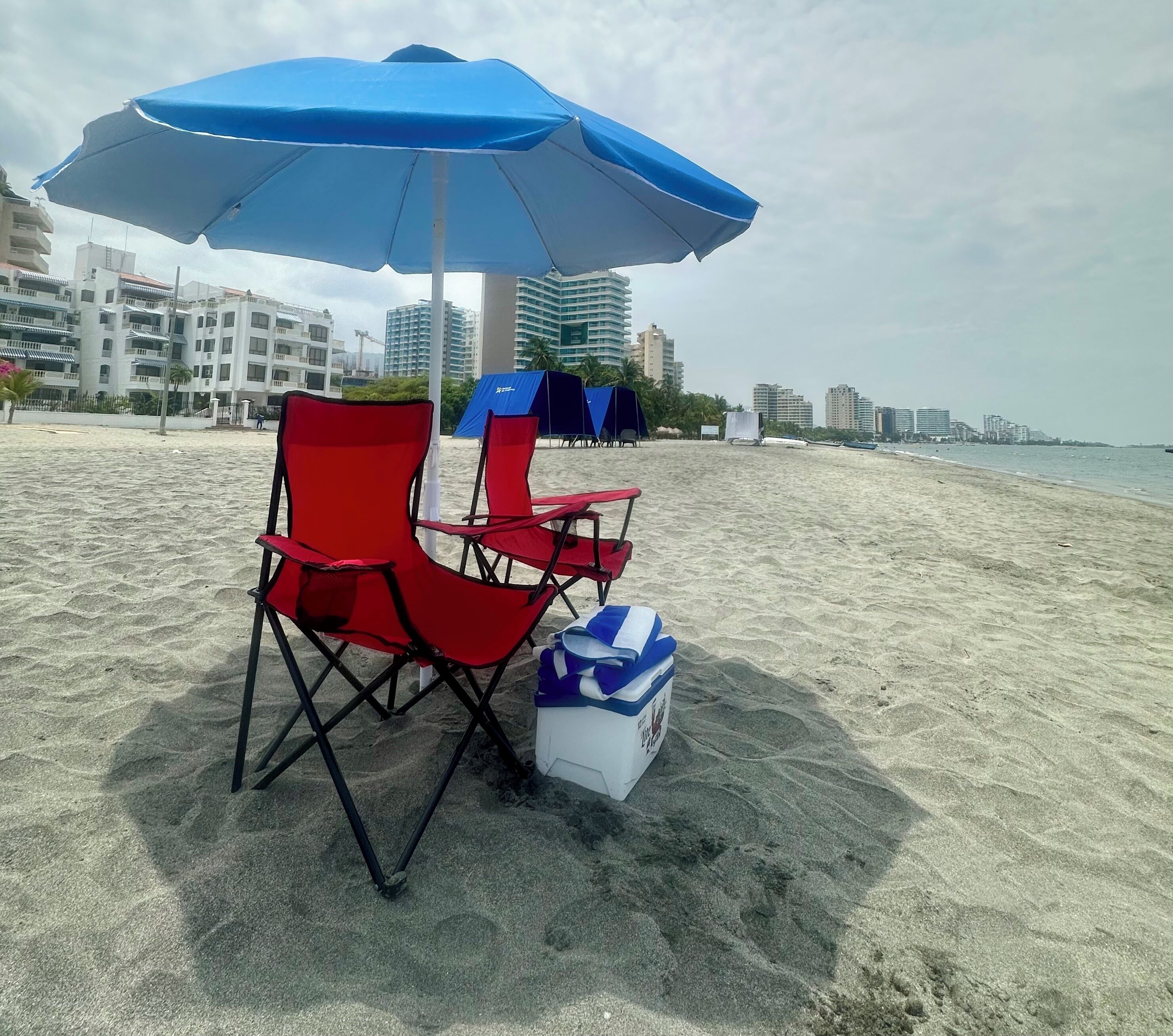 A beach with a blue umbrella, red chairs, and a cooler on the sand.