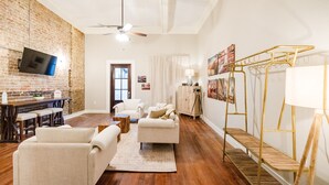 Ein Wohnzimmer mit Backsteinwand, Deckenventilator, Flachbildfernseher, Essbereich mit Tisch und Barhocker sowie eine Treppe mit goldener Geländer.