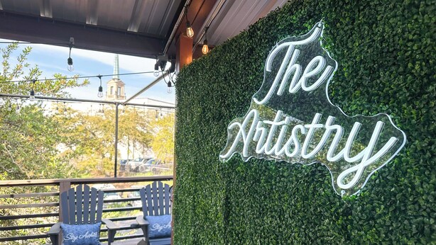 Ein Neon-Schild mit der Aufschrift 'The Artistry' vor dem Hintergrund von dichtem Grünwerk.