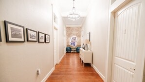Un couloir avec des tableaux encadrés accrochés au mur, un mur d’accent en briques, un lustre, un coin salon avec des chaises et une table, et un meuble blanc.