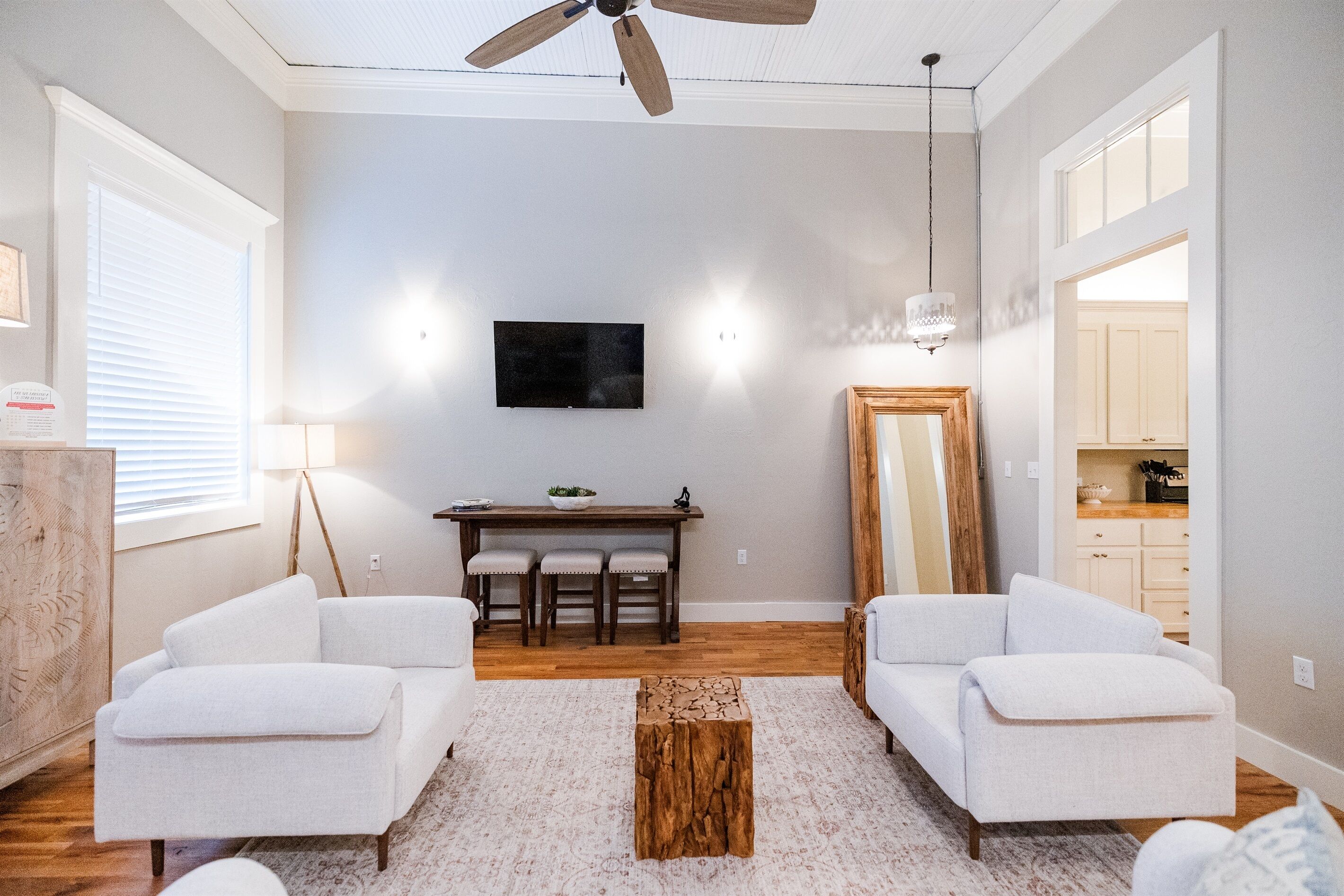 Un salon moderne doté d’un ventilateur de plafond, d’un téléviseur à écran plat fixé au mur, d’un coin repas avec une table et des chaises en bois, et d’un grand miroir.