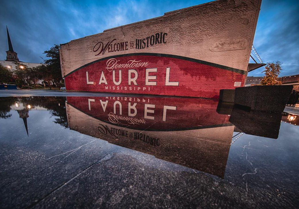 Placa de boas-vindas para o centro histórico de Laurel, Mississippi, com reflexo nítido no pavimento molhado.