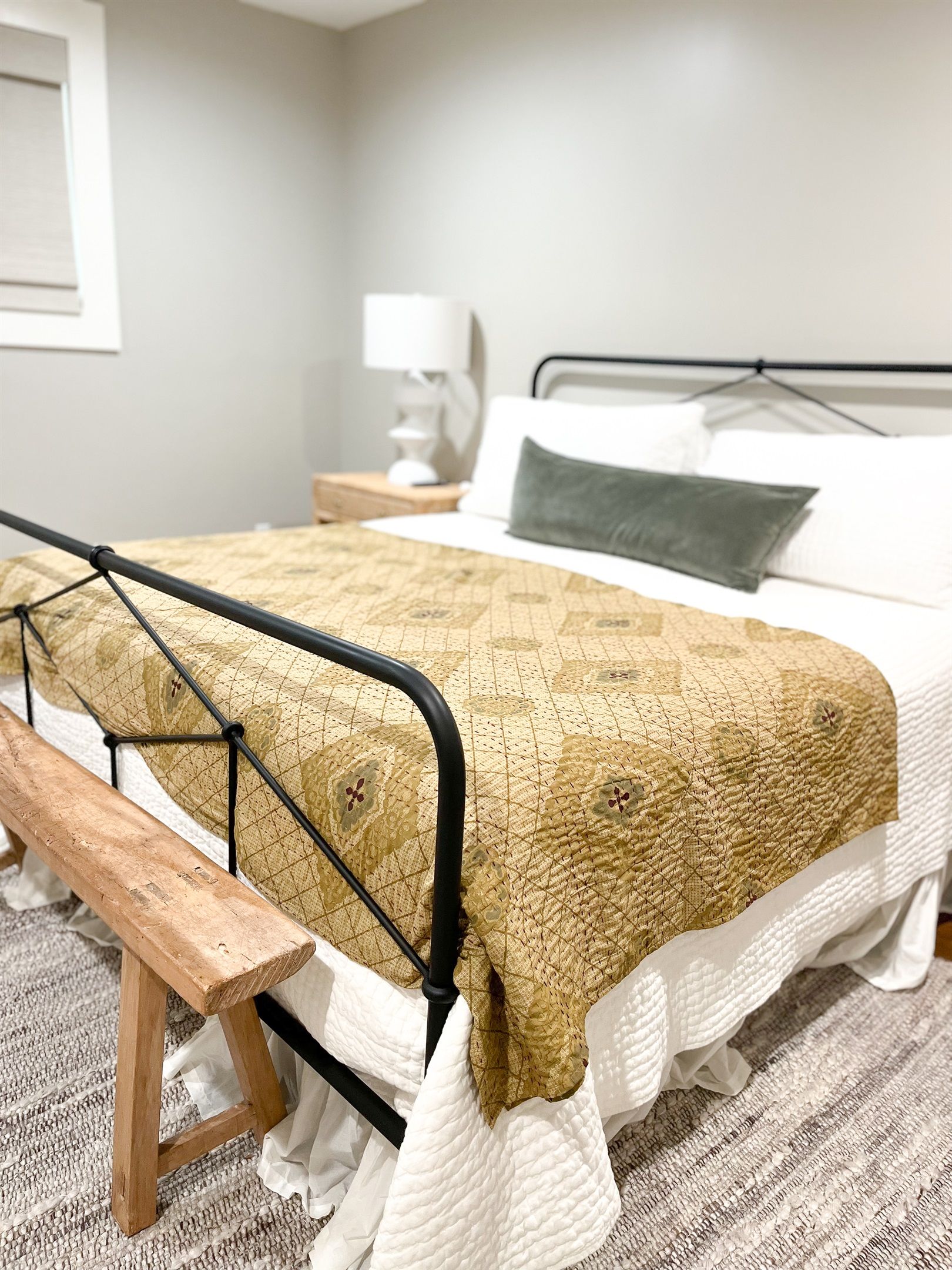 A bed with a metal frame, a wooden bench, a patterned blanket, and a bedside table with a lamp.