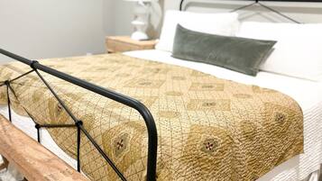 A bed with a metal frame, a wooden bench, a patterned blanket, and a bedside table with a lamp.