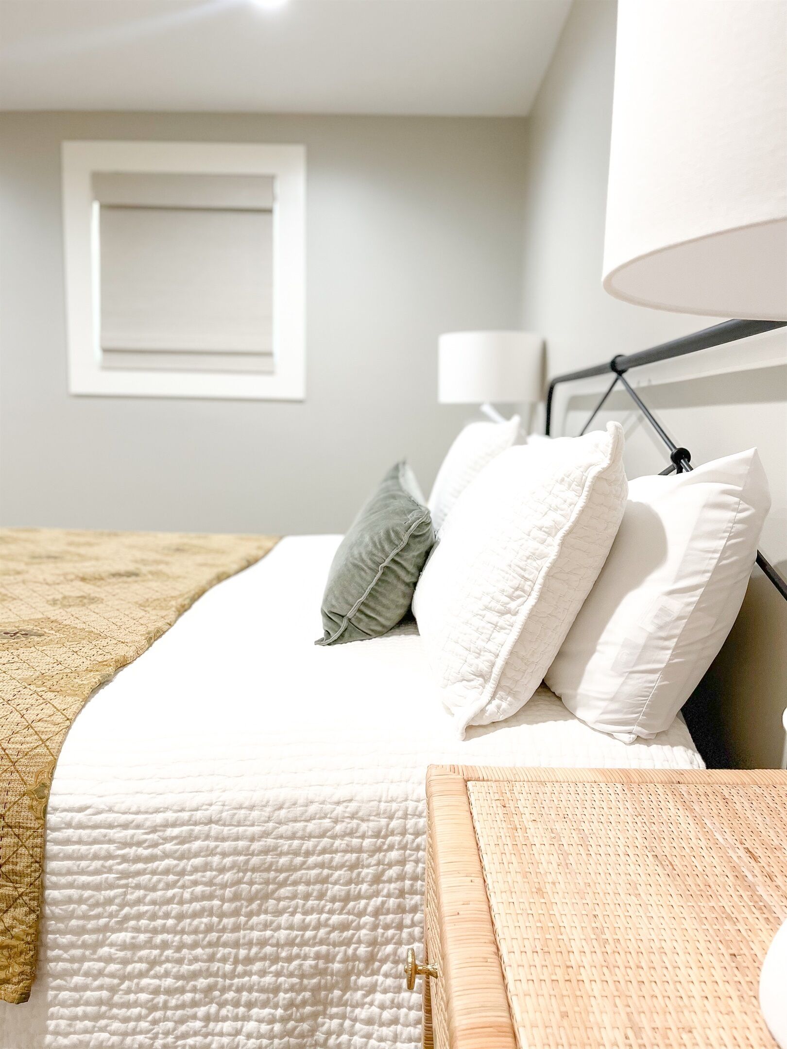Una cama bien hecha con ropa de cama blanca, un almohadón gris y una mesita de noche de madera con una lámpara.