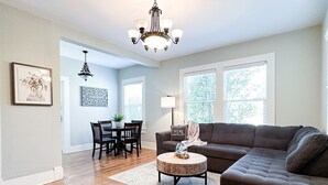 A spacious living room with hardwood flooring, a dark brown sectional sofa, a round coffee table, and a chandelier. There is a dining area with a table and chairs visible in the background. - Centrally Located Downtown, Full Kitchen, Patio (Laurel)