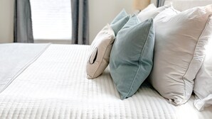 Un lit bien fait, recouvert d’une couverture blanche à texture intéressante, avec des rideaux gris et une lampe sur une table de chevet.