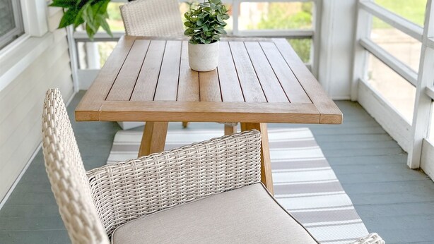 Een overdekt terras met een houten tafel en rieten stoelen, een plant in pot, en een gebouw op de achtergrond.