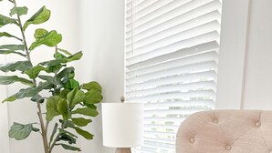 Een woonkamer met een beige bank, een kleine potplant, een houten bijzettafeltje met een lamp en een raam met zonwering.