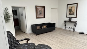 A living room with a dark leather sofa, a black leather armchair, and a wooden coffee table. There are two framed artworks on the wall and a potted plant on the left. - THE PLACE (Las Vegas)