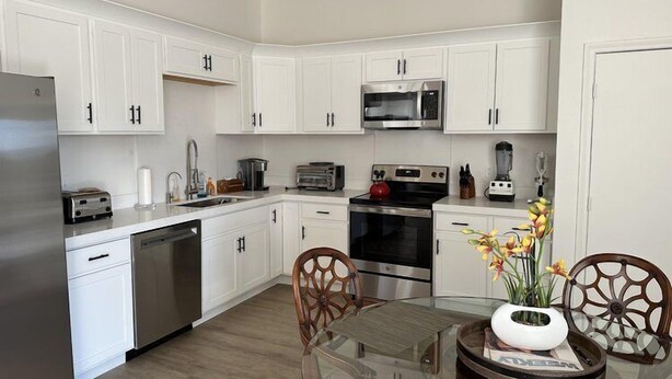 A modern kitchen with white cabinets, stainless steel appliances, and a glass dining table. - THE PLACE (Las Vegas)