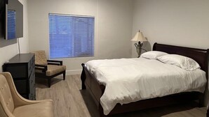A bedroom with a bed, a desk, a chair, a lamp, and a window with blinds. - THE PLACE (Las Vegas)