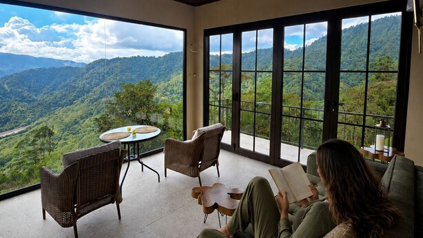 Una habitación con un ventanal grande, un ventilador de techo y una persona leyendo un libro.