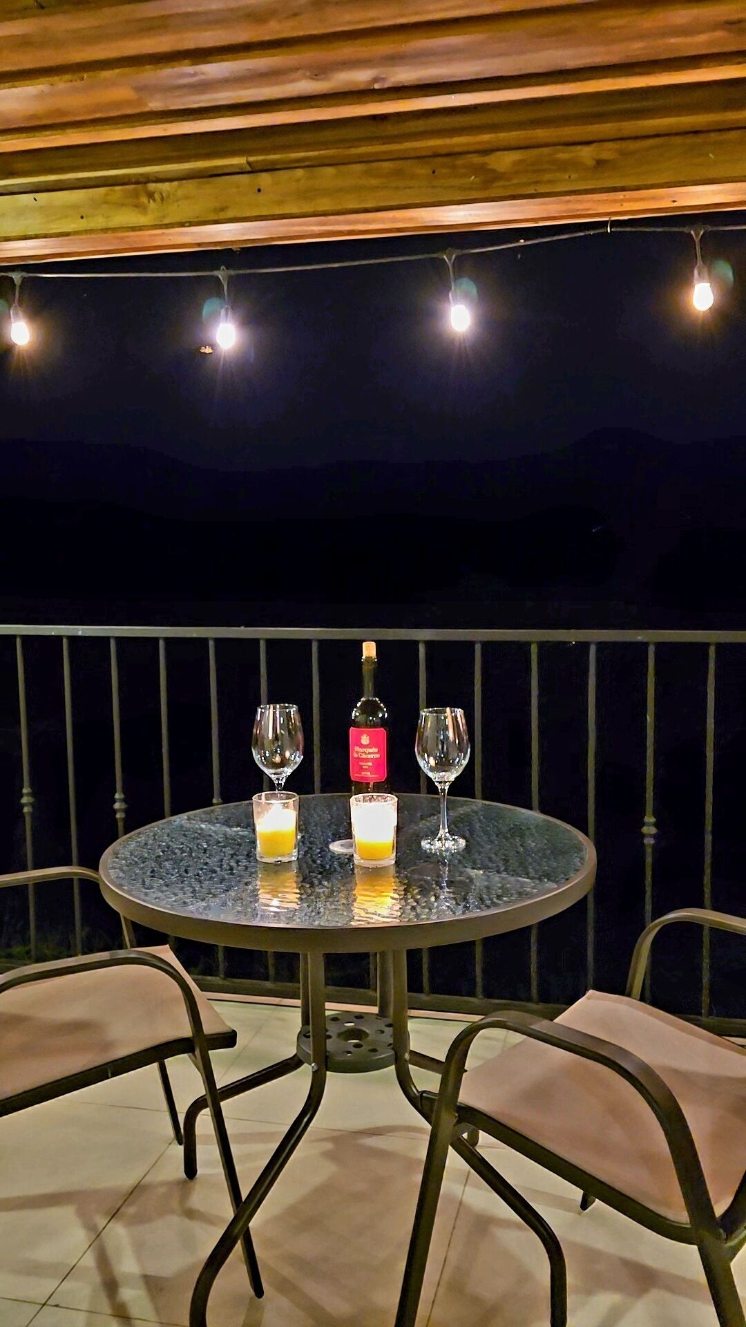A round table with two glasses and a bottle of wine, surrounded by chairs, on an outdoor balcony with a railing and string lights overhead.
