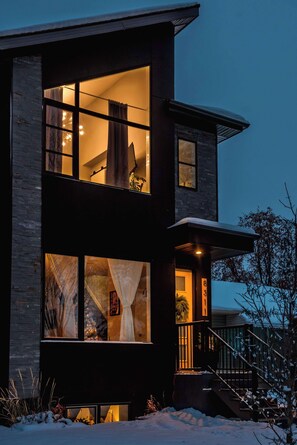 Una casa moderna de dos pisos con ventanas amplias, una escalera visible y un terreno cubierto de nieve.