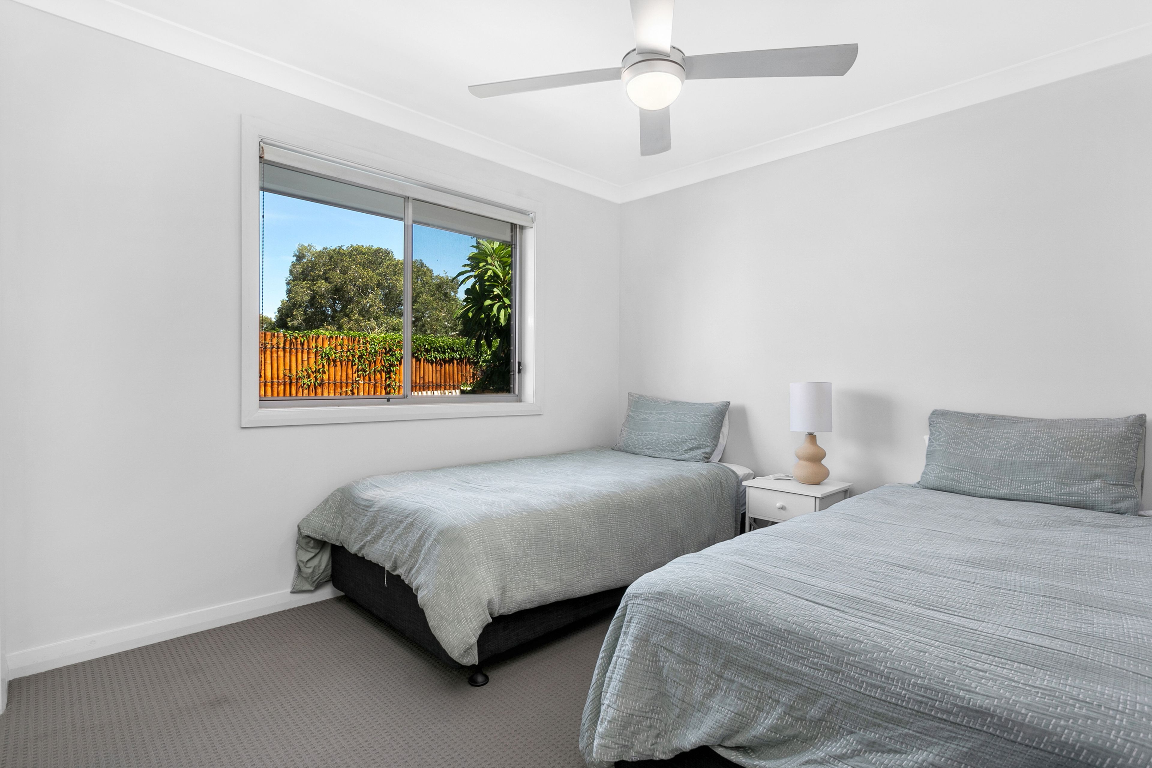 A bedroom with two beds, a ceiling fan, a window with a view of a fence and trees, and a bedside table with a lamp.