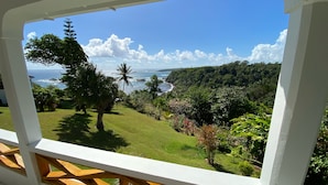 Une vue depuis un balcon doté d’une balustrade en bois, donnant sur un paysage verdoyant et luxuriant, un palmier et une côte bordée par l’océan et le ciel.