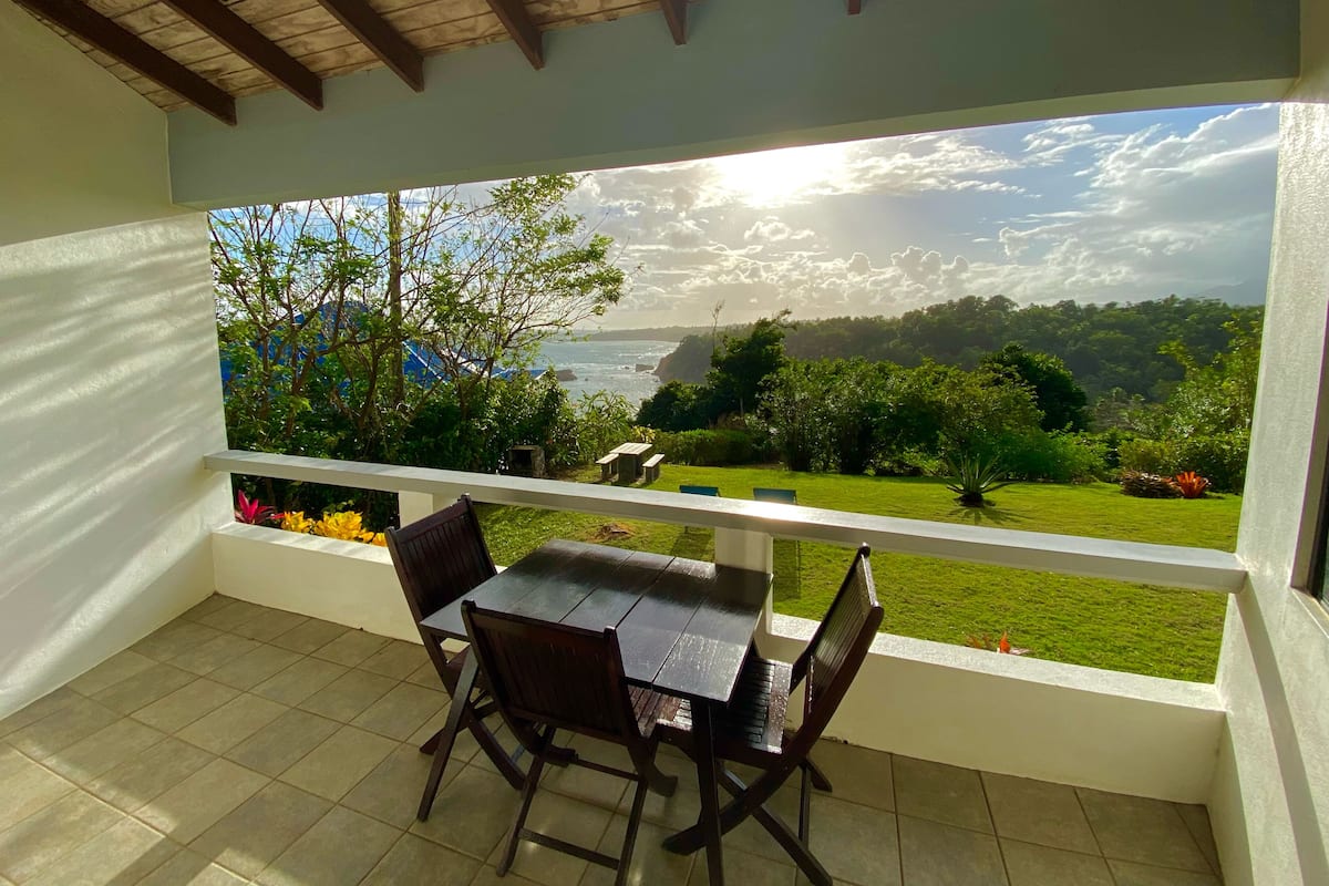 A covered patio with a table and chairs, a view of a lush landscape, and a wooden ceiling.
