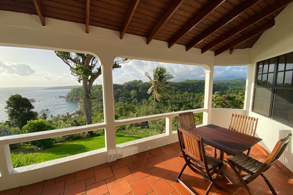 A covered patio with a table and chairs, offering a view of a coastal landscape with greenery and a body of water.
