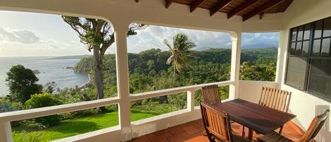 Um alpendre coberto com mesa e cadeiras, oferecendo vista para uma paisagem costeira com vegetação e um corpo d'água.