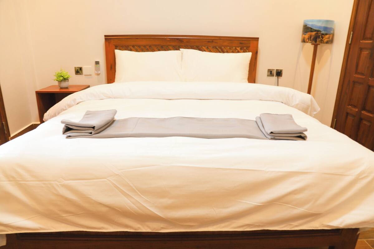 A neatly made bed with white linens and folded towels, a wooden headboard, a bedside table with a plant, and a painting on the wall.