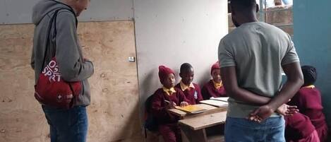 A classroom with students seated at desks, a teacher standing at the front, and a man standing at the back.