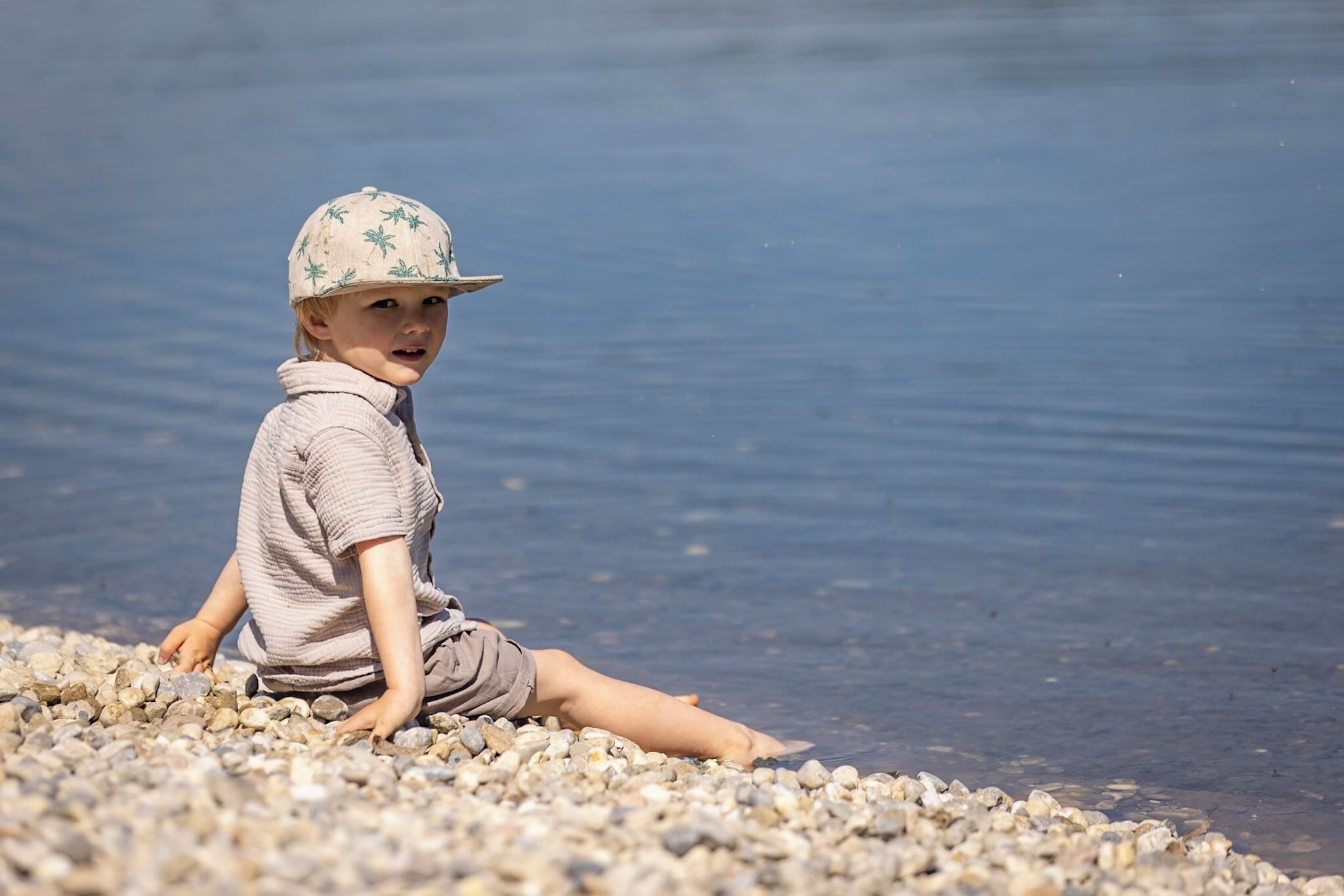 Niño sentado en una playa pedregosa cerca de aguas tranquilas.