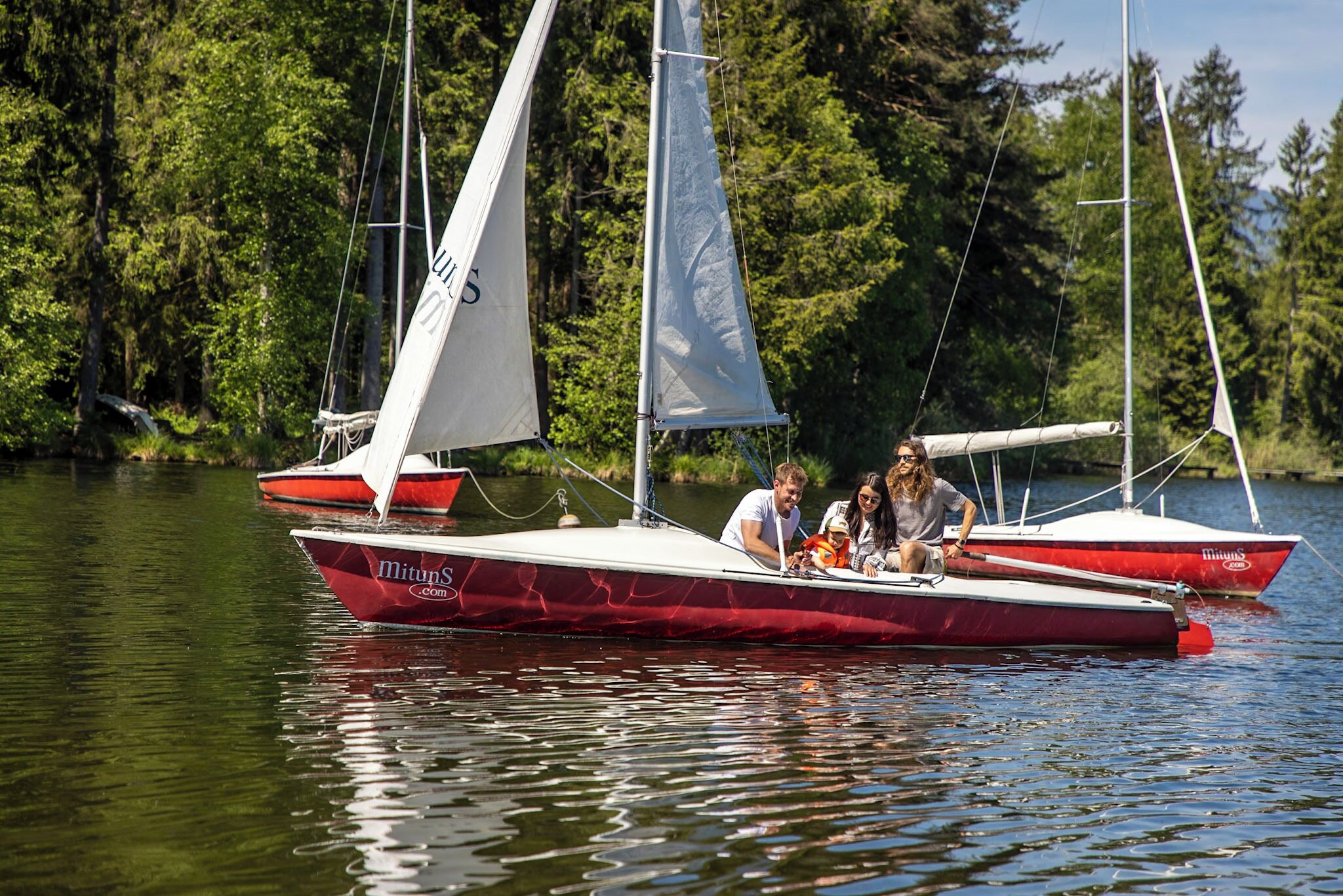 Un velero rojo con cabina blanca y una persona timoneando, rodeado de agua tranquila y árboles.