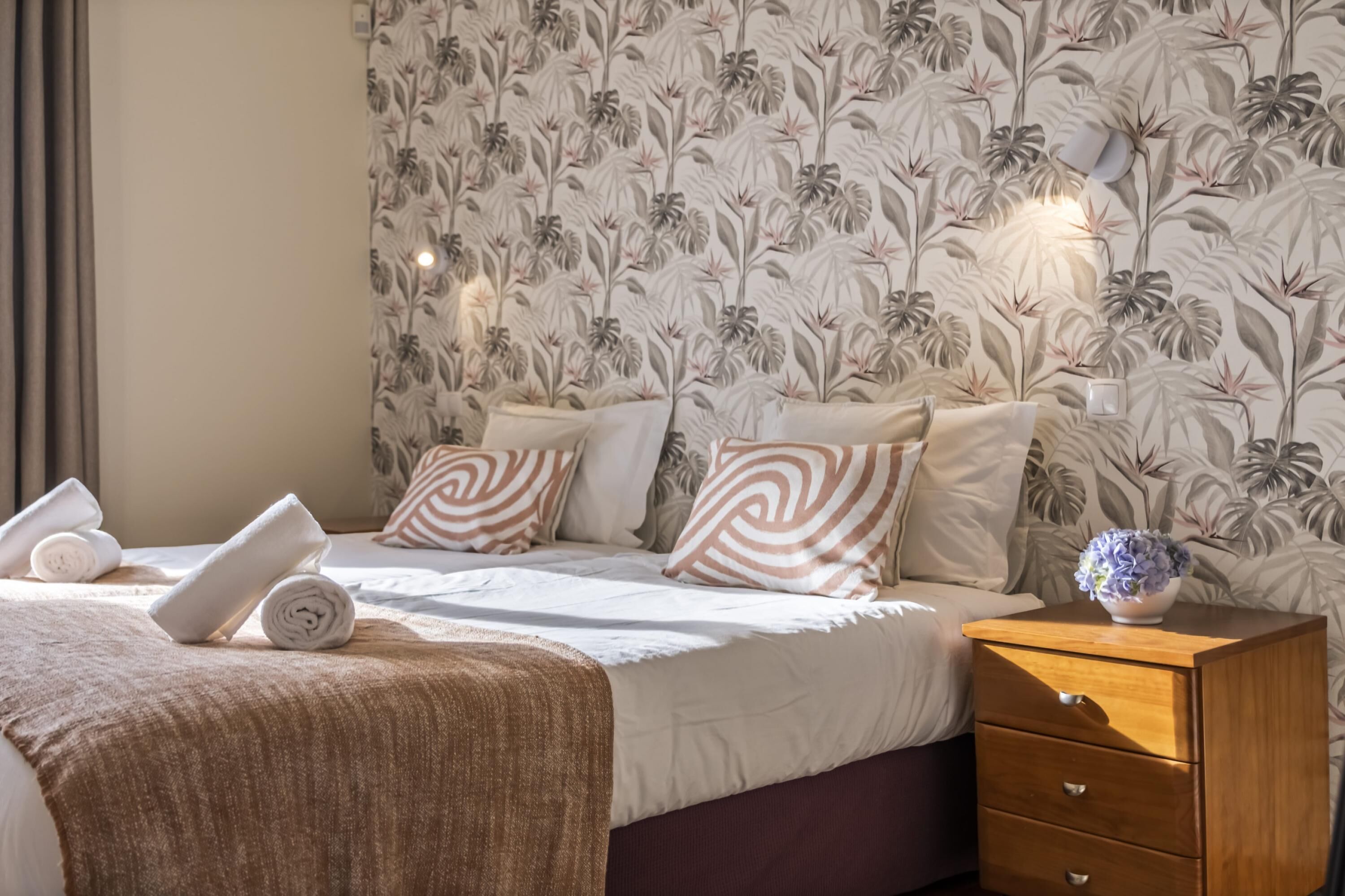 A neatly made bed with a brown blanket, a wooden nightstand with a vase, and a floral wallpaper pattern.