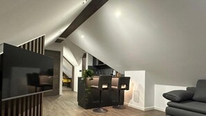 A modern living room with a black sofa, a black ottoman, and a wooden floor. There is a kitchen area with a bar and stools, and a staircase leading to an upper level. - 4 Elements Resort Bled Studio with sofa bed (Radovljica)