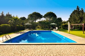 A rectangular swimming pool with a stone deck surrounding it, set in a landscaped garden with trees and umbrellas.