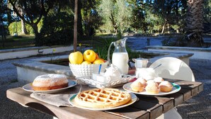 A picnic table with a variety of food items including a cake, waffles, muffins, and a pitcher of milk, set in an outdoor garden area.